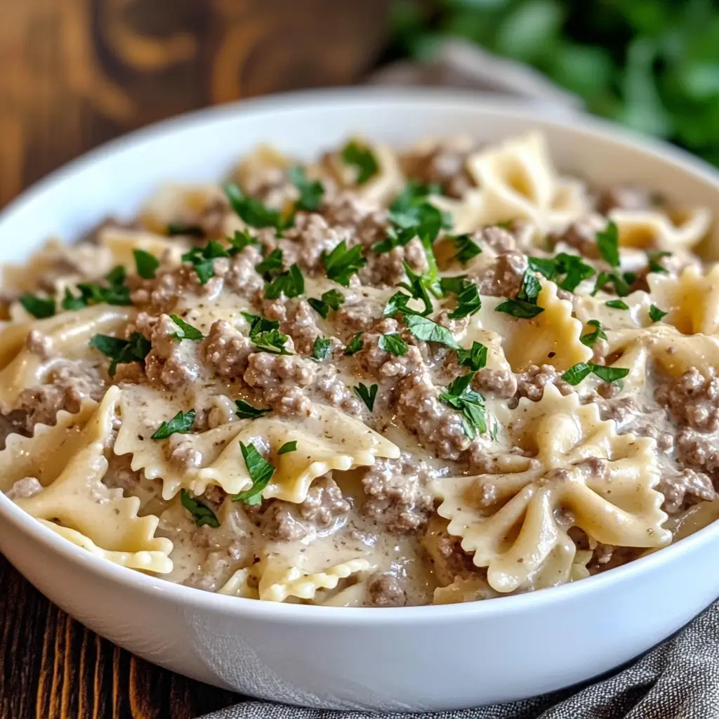 Homemade creamy beef and bowtie pasta in a white bowl, garnished with freshly chopped parsley. The dish features tender farfalle pasta coated in a rich Alfredo sauce with seasoned ground beef.